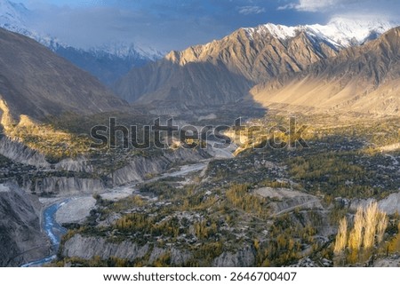 A clear blue river flows through a wide valley, surrounded by green trees and tall, rocky mountains. Small houses rest among the hills, touched by soft sunlight