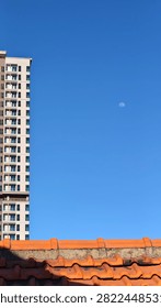 Clear blue sky frames a tall building. Orange tiles contrast with the bright sky. Moon hangs low, adding quiet beauty. Urban and natural elements blend harmoniously