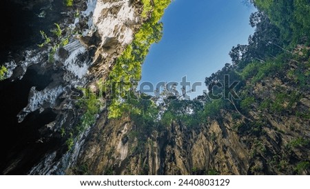 The clear blue sky is visible in a frame formed by sheer cliffs. Green tropical vegetation on steep rocky slopes. Bottom view. Malaysia. Batu caves. Kuala Lumpur