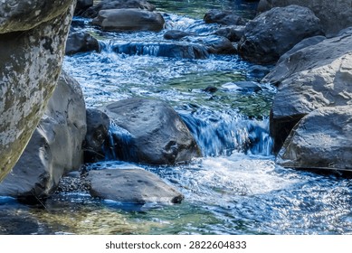 Clear blue stream cascading down small rock steps, creating a mini waterfall surrounded by large boulders.