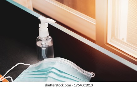 A Clear Bottle Of Alcohol Gel On The Table, A Protective Mask Placed Next To It, And Orange Sunlight Shines Through The Window.