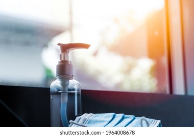 A Clear Bottle Of Alcohol Gel On The Table, A Protective Mask Placed Next To It, And Orange Sunlight Shines Through The Window.