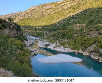 A clear, calm river flows through a rocky canyon in Montenegro, with green hills and bright sunlight reflecting perfectly on the water surface.