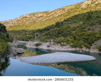 A clear, calm river flows through a rocky canyon in Montenegro, with green hills and bright sunlight reflecting perfectly on the water surface.