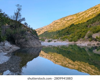 A clear, calm river flows through a rocky canyon in Montenegro, with green hills and bright sunlight reflecting perfectly on the water surface.