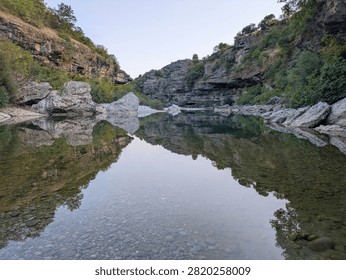 A clear, calm river flows through a rocky canyon in Montenegro, with green hills and bright sunlight reflecting perfectly on the water surface.