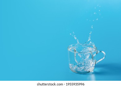 Clear cold water in a glass cup on a blue background. Beautiful splash from falling ice