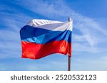 A clear daytime view of the Russian national flag waving in the wind against a blue sky.