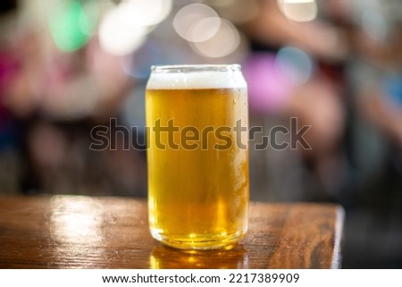 A clear drinking glass in the shape of a beer can filled with cold froth lager ale. The Belgian pint sits on the edge of a wooden patio table at a microbrewery. The background has multicolored lights.
