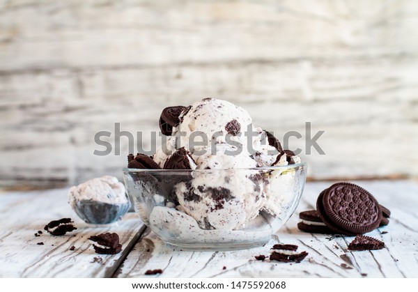 Clear glass bowl of cookies and\
cream ice cream. Selective focus with blurred background. Whole and\
crumbled cream filled cookies scattered about table.\
