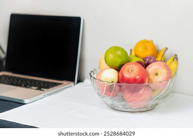 A clear glass bowl filled with various fruits including apples, bananas, a lime, a pear, and a plum, placed on a white tablecloth next to a laptop. - Powered by Shutterstock