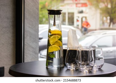 A clear glass carafe, jug of lemonade with lemons, mint next to glasses on a round black table in a cafe or restaurant against a panoramic window. A refreshing cold summer citrus drink. Iced punch.