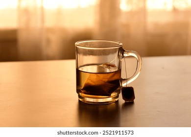 A clear glass mug filled with brewed tea, with a partially submerged tea bag visible, sits on a wooden table, bathed in warm sunlight streaming through a nearby window - Powered by Shutterstock