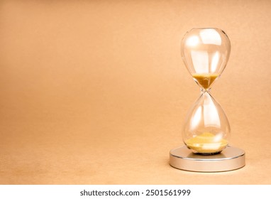 A clear hourglass with gold sand on chrome podium on light brown recycled paper background with copy space. Golden sand in sandglass clock isolated on eco paper. Time, timer countdown, deadline watch.