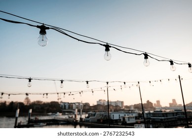 Clear lightbulbs hanging on string lights across an outdoor entertainment area, with a blurred city skyline and river in the background during twilight