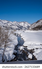 A clear mountain stream flows between snow-covered banks, surrounded by frosty trees and snowy peaks under a bright blue sky. A peaceful winter landscape showcasing nature's pure, icy beauty.