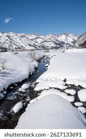 A clear mountain stream flows between snow-covered banks, surrounded by frosty trees and snowy peaks under a bright blue sky. A peaceful winter landscape showcasing nature's pure, icy beauty.