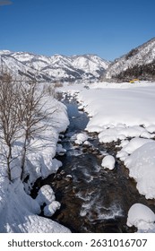 A clear mountain stream flows between snow-covered banks, surrounded by frosty trees and snowy peaks under a bright blue sky. A peaceful winter landscape showcasing nature's pure, icy beauty.