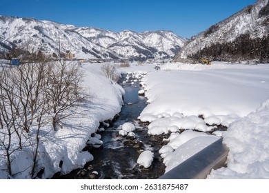 A clear mountain stream flows between snow-covered banks, surrounded by frosty trees and snowy peaks under a bright blue sky. A peaceful winter landscape showcasing nature's pure, icy beauty.