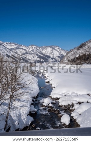 A clear mountain stream flows between snow-covered banks, surrounded by frosty trees and snowy peaks under a bright blue sky. A peaceful winter landscape showcasing nature's pure, icy beauty.