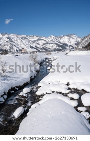A clear mountain stream flows between snow-covered banks, surrounded by frosty trees and snowy peaks under a bright blue sky. A peaceful winter landscape showcasing nature's pure, icy beauty.