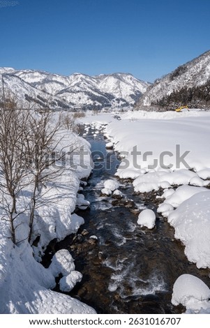 A clear mountain stream flows between snow-covered banks, surrounded by frosty trees and snowy peaks under a bright blue sky. A peaceful winter landscape showcasing nature's pure, icy beauty.