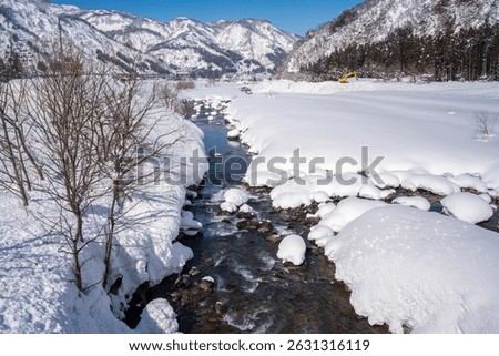 A clear mountain stream flows between snow-covered banks, surrounded by frosty trees and snowy peaks under a bright blue sky. A peaceful winter landscape showcasing nature's pure, icy beauty.