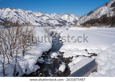 A clear mountain stream flows between snow-covered banks, surrounded by frosty trees and snowy peaks under a bright blue sky. A peaceful winter landscape showcasing nature's pure, icy beauty.