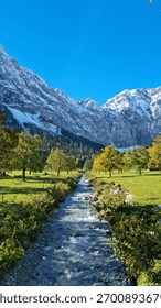 A clear mountain stream flows through a green valley surrounded by autumn trees and snow-covered peaks under a bright blue sky