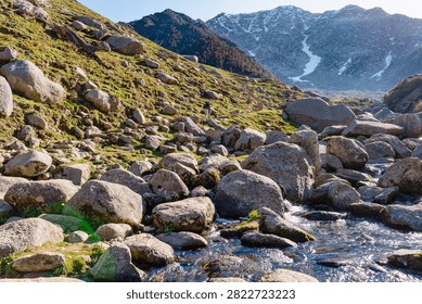 A clear mountain stream flows through a rocky alpine landscape with snow-capped peaks in the background.