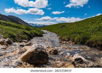 A Clear Mountain Stream Winds Through Alpine Meadows Toward Distant Snow-Covered Peaks - Crystal-clear water flows over smooth stones and small rocks through an alpine meadow surrounded by rolling green hills. The stream creates a natural leading line toward a distant mountain range marked by patches of snow on the highest summits. Bright sunlight reflects from the moving water, highlighting its clarity and texture, while scattered clouds add depth to the blue summer sky above. The photograph captures the freshness and vitality of a high-mountain landscape shaped by snowmelt, flowing water, and seasonal vegetation.
