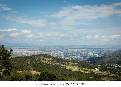 Clear mountain view of Tbilisi, Georgia, with rolling hills in the foreground and a partly cloudy sky above. The city stretches into the distance, framed by a scenic horizon - Powered by Shutterstock