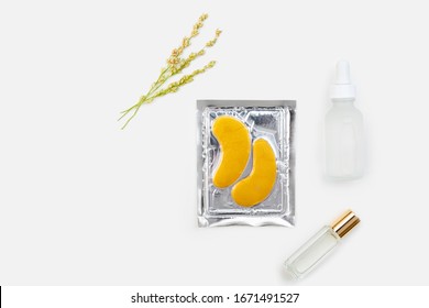 Clear Perfume, White Glass Eye Dropper Serum Bottle, And Gold Collagen Eye Masks Arranged With Golden Wildflowers / Still Life Isolated On Gray Surface