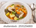 Clear Pike soup with vegetables, parsley and spices in white bowl on white wooden table with spoon, close-up