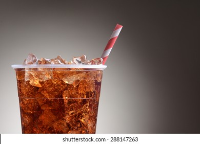 A Clear Plastic Cup Filled With Ice And Cola. Only Half The Cup Is Shown With A Red And White Striped Straw Over A Light To Dark Gray Background.