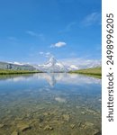 Clear reflection of the Matterhorn mountain in a tranquil alpine lake, surrounded by lush greenery and snow-capped peaks under a bright blue sky, capturing the serene beauty of the Swiss Alps