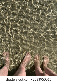 A clear shallow ocean looking down to a sandy beach and two pairs of feet. 