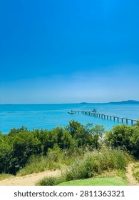 A clear sky shows no clouds as the sunlight shines over the blue sea. The long pier reaches out into the water, and green trees are visible along the coast.