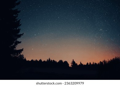 Clear starry night sky with a radiant orange hue on the horizon following sunset. The serene cosmic vista is elegantly framed by the silhouette of trees at the bottom and left