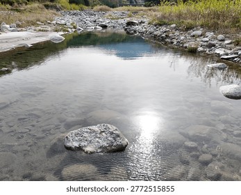 Clear stream flows gently over smooth stones. Ripples catch sunlight, glimmering softly. Rocks rest peacefully in shallow water. Bankside grasses sway in subtle breeze