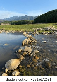 Clear stream flows over smooth rocks with green meadows and forested mountains in the distance