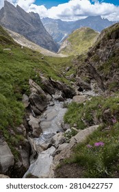 A clear stream flows through a lush, verdant alpine gorge, framed by dramatic European mountain peaks under a bright, dynamic cloudy sky.