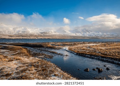 A clear stream flows towards a large fjord in Iceland. Majestic snow-covered mountains stand out in the background under a blue, cloudy sky. The surrounding ground shows shades of dry grass, rocks, and light patches of winter snow.