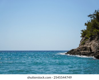 Clear turquoise waters meet a rugged rocky coastline lined with pine trees under a cloudless blue sky, showcasing the peaceful and untouched beauty of a secluded seascape. - Powered by Shutterstock