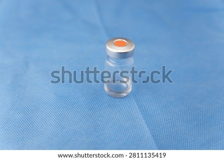 Clear vial with silver cap sits on blue surgical cloth. Cap features orange seal for secure closure. Medical or lab settings imply sterile conditions