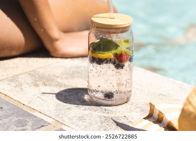 A clear water bottle with lemon and berries sits by a poolside. Sunlight reflects off the water, suggesting a warm, leisurely day outdoors. - Powered by Shutterstock