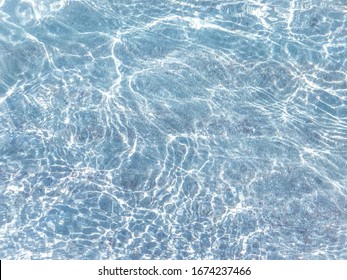 Clear Water In The Sea, Sun Glare, Waves And Sea Pebbles. Light Blue Sea Water Background