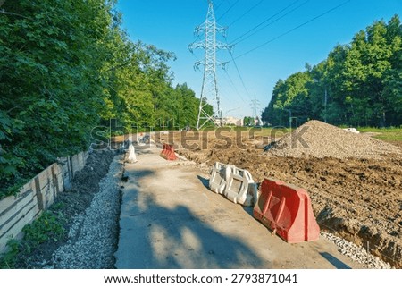 A cleared utility corridor cutting through a green forest with high-voltage steel power pylons and overhead lines. A freshly laid dirt and gravel pathway runs