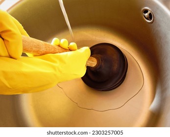 Clearing the blockage using a plunger. Clogged pipes and kitchen sink. Plumbing and maintenance concept.Yellow rubber gloves and manual plunger in bathroom 