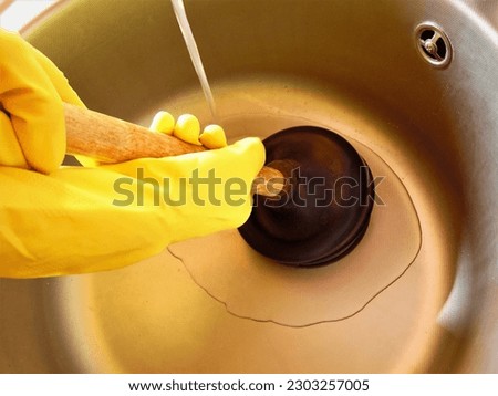 Clearing the blockage using a plunger. Clogged pipes and kitchen sink. Plumbing and maintenance concept.Yellow rubber gloves and manual plunger in bathroom 